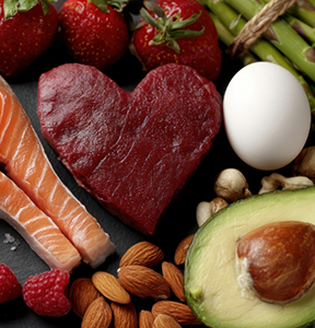 Piece of meat cut into a heart shape next to nuts, avocados, salmon, and other healthy foods